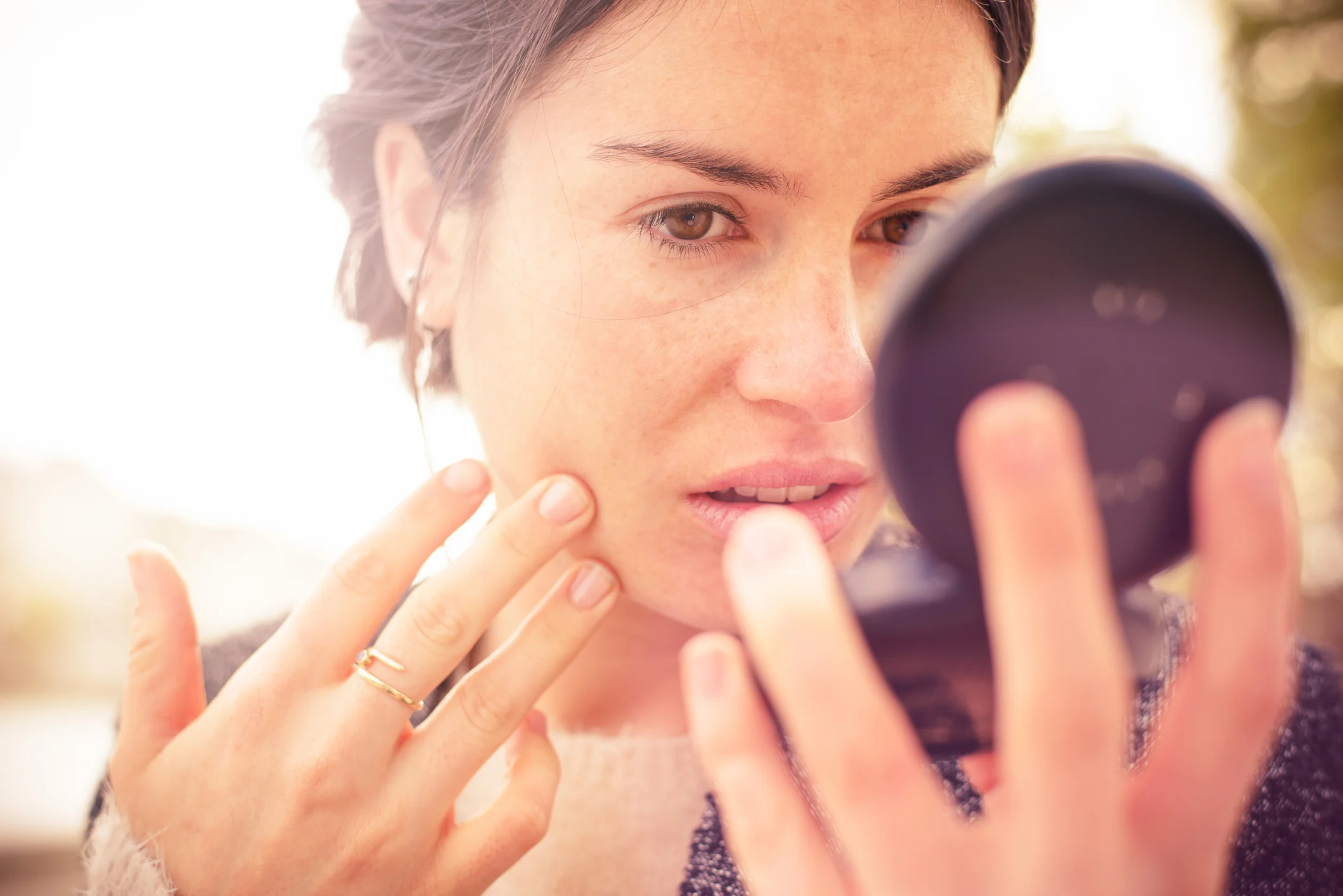 Young woman outdoors checking her face in a round powder compact mirror.