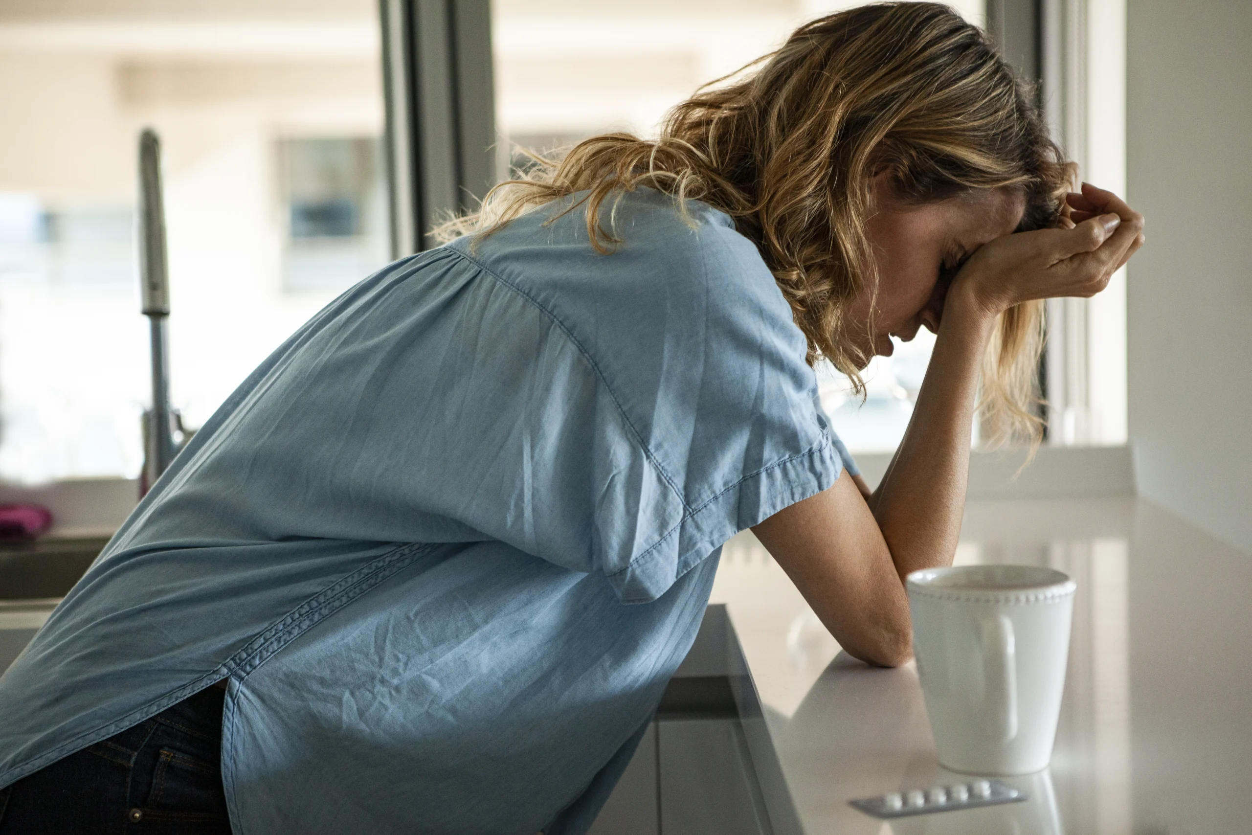portrait of woman bent over counter with hangover with cup of coffee sitting on counter next to her